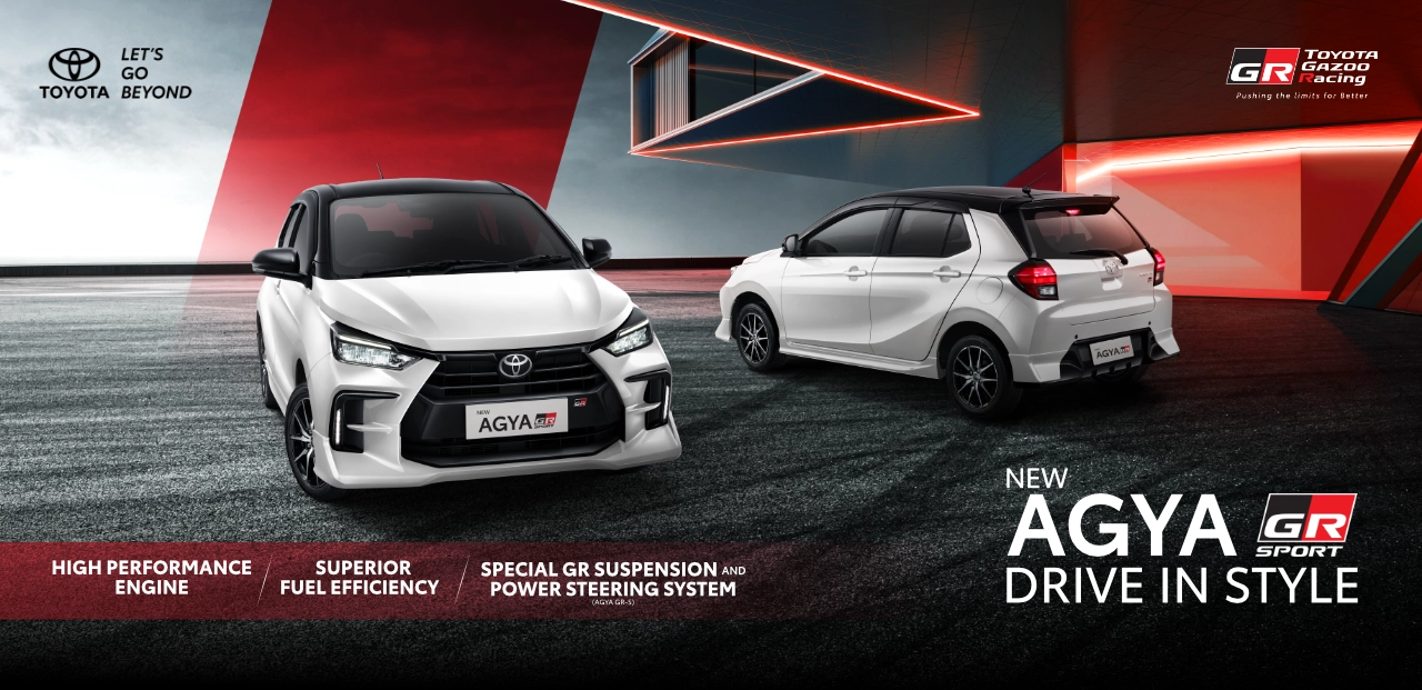 Dua unit mobil hatchback Toyota New Agya GR Sport warna putih dengan atap hitam (two-tone) tampak depan dan belakang di area berdesain modern dengan aksen cahaya merah. Banner Tunas Toyota ini menampilkan slogan Drive in Style serta menonjolkan fitur mesin performa tinggi dan suspensi khusus GR