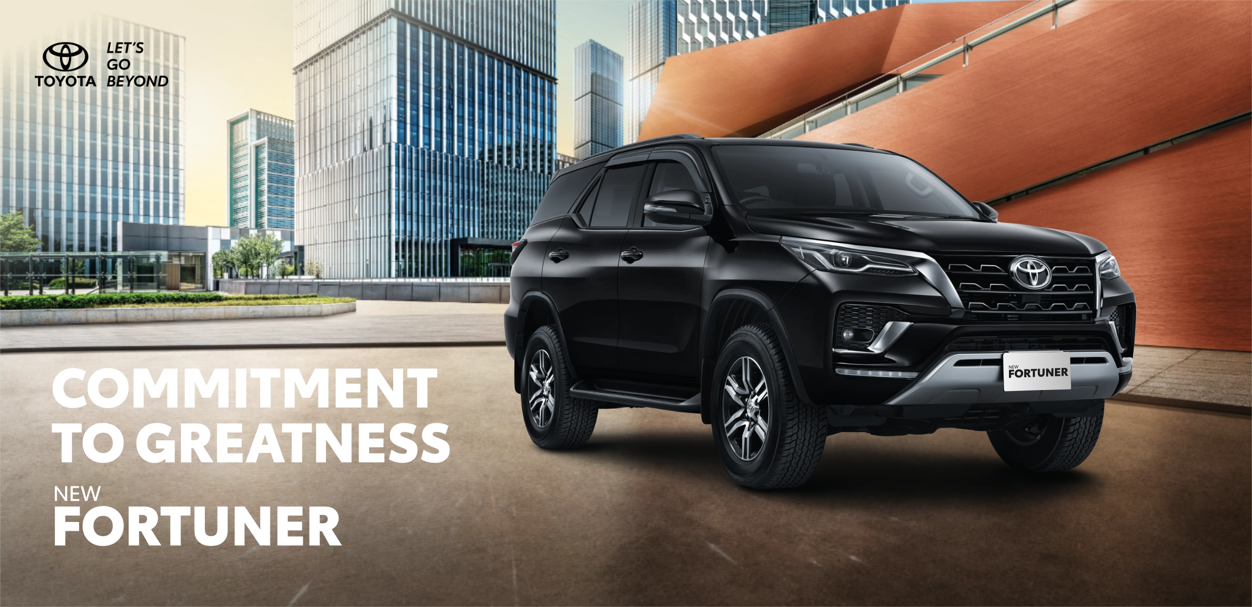 Banner promosi Toyota New Fortuner warna hitam yang tampil elegan dan gagah di tengah lanskap gedung-gedung perkotaan modern, memuat tagline Commitment to Greatness.