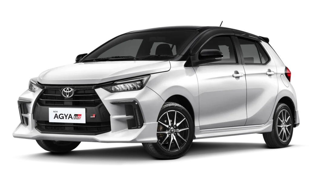 Tampilan studio tampak depan samping (3/4) mobil hatchback kompak Toyota New Agya GR Sport warna putih dengan atap dan spion berwarna hitam (two-tone). Mobil ini memiliki latar belakang hitam pekat yang menonjolkan body kit agresif, gril dengan emblem GR, dan velg alloy sporty. Gambar produk utama resmi dari Tunas Toyota